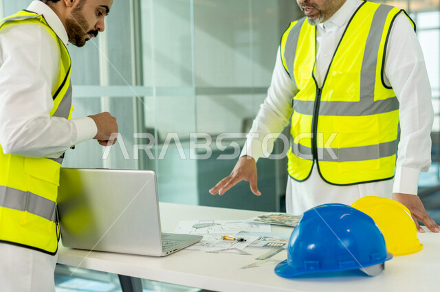 إجتماع مهندسان معماريان سعوديان خليجيان يعملان في مجال الهندسة المعماري في داخل المكتب في المملكة العربية السعودية, إرتداء سترة الحماية الخاصة بالعمل, تدقيق العمل، الإشراف والنقاش، مفهوم الهندسة، مهن ووظائف هندسية ، فريق عمل خبير، خطط ومشاريع معمارية