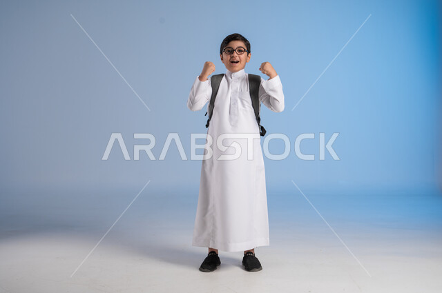 المدارس الأكاديمية في المملكة العربية السعودية, الاستعداد للعام الدراسي الجديد،  تلميذ عربي خليجي سعودي يرتدي الثوب التقليدي والنظارات الطبية يحمل حقيبة الظهر المدرسية يرفع يديه للأعلى، ايماءات الحماس والسعادة بالعودة إلى المدرسة، صورة الجسم بالطول الكامل، خلفية زرقاء