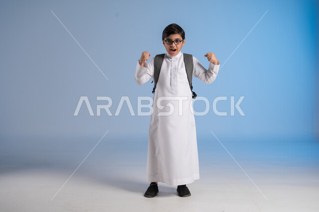 المدارس الأكاديمية في المملكة العربية السعودية, الاستعداد للعام الدراسي الجديد،  تلميذ عربي خليجي سعودي يرتدي الثوب التقليدي والنظارات الطبية يحمل حقيبة الظهر المدرسية يرفع يديه للأعلى، ايماءات الحماس والسعادة بالعودة إلى المدرسة، صورة الجسم بالطول الكامل، خلفية زرقاء
