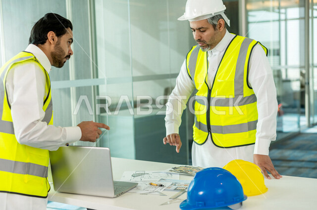 إجتماع مهندسان معماريان سعوديان خليجيان يعملان في مجال الهندسة المعماري في داخل المكتب في المملكة العربية السعودية, إرتداء سترة الحماية الخاصة بالعمل, تدقيق العمل، الإشراف والنقاش، مفهوم الهندسة، مهن ووظائف هندسية ، فريق عمل خبير، خطط ومشاريع معمارية