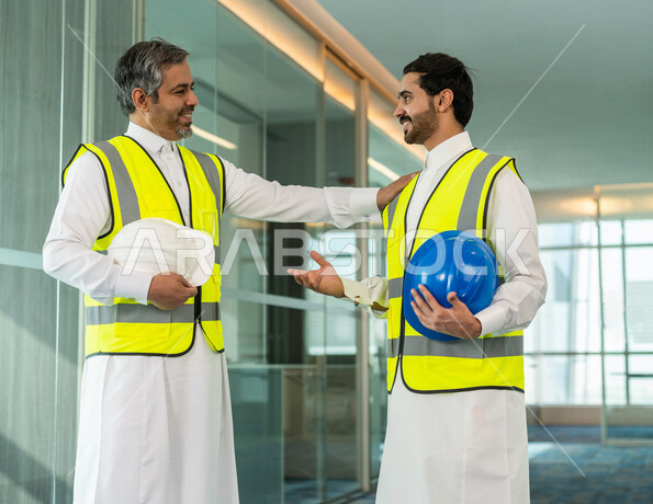 A meeting of a Saudi engineer and a Saudi worker inside the company in Saudi Arabia, discussing to start a new project, working in the field of engineering, wearing a work protection jacket, auditing work, supervision and discussion, engineering professio
