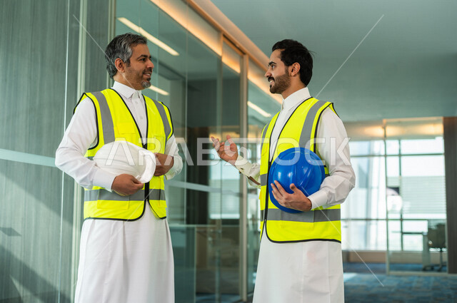 إجتماع مهندس سعودي وعامل سعودي في داخل الشركة بالمملكة العربية السعودية، يتناقشان لبدء مشروع جديد، العمل في مجال الهندسة, إرتداء سترة الحماية الخاصة بالعمل, تدقيق العمل، الإشراف والنقاش، مهن ووظائف هندسية، فريق عمل خبير، خطط ومشاريع معمارية