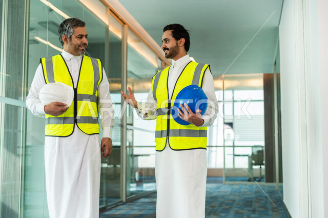 A meeting of a Saudi engineer and a Saudi worker inside the company in Saudi Arabia, discussing to start a new project, working in the field of engineering, wearing a work protection jacket, auditing work, supervision and discussion, engineering professio