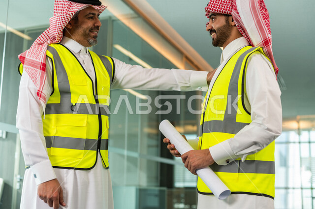 مهندسان معماريان سعوديان خليجيان يحملان مخطط لمشروع البناء للتخطيط وتنفيذ العمل في داخل المكتب في المملكة العربية السعودية, إرتداء سترة الحماية الخاصة بالعمل, تدقيق العمل، الإشراف والنقاش في العمل، مفهوم الهندسة، مهن ووظائف هندسية