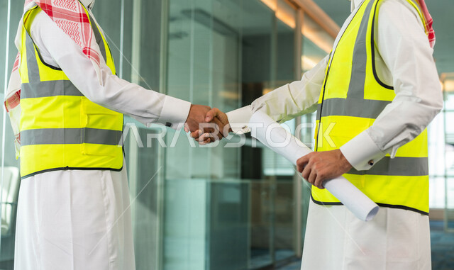 إجتماع مهندسان معماريان سعوديان خليجيان ، يتصافحان لبدء مشروع جديد، العمل في مجال الهندسة في داخل المكتب في المملكة العربية السعودية, إرتداء سترة الحماية الخاصة بالعمل, تدقيق العمل، الإشراف والنقاش، مهن ووظائف هندسية، فريق عمل خبير، خطط ومشاريع معمارية