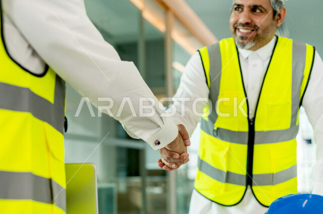 إجتماع مهندسان معماريان سعوديان خليجيان ، يتصافحان لبدء مشروع جديد، العمل في مجال الهندسة في داخل المكتب في المملكة العربية السعودية, إرتداء سترة الحماية الخاصة بالعمل, تدقيق العمل، الإشراف والنقاش، مهن ووظائف هندسية، فريق عمل خبير، خطط ومشاريع معمارية