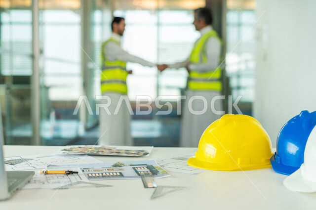 إجتماع مهندسان معماريان سعوديان خليجيان ، يتصافحان لبدء مشروع جديد، العمل في مجال الهندسة في داخل المكتب في المملكة العربية السعودية, إرتداء سترة الحماية الخاصة بالعمل, تدقيق العمل، الإشراف والنقاش، مهن ووظائف هندسية، فريق عمل خبير، خطط ومشاريع معمارية