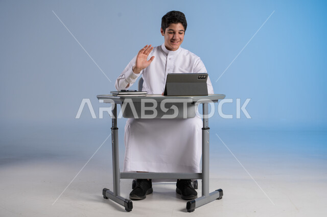 تلقي دروس الاون لاين عبر الانتلانت، مفهوم العودة إلى المدارس، طرائق تعليمية جديدة متطورة، بورتريه لطالب عربي خليجي سعودي يرتدي الثوب التقليدي يجلس في مقعده بايماءات السعادة، دمج التكنولوجيا بالتعليم في المدارس الحديثة، خلفية زرقاء