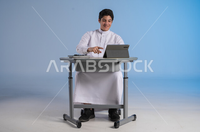 تلقي دروس الاون لاين عبر الانتلانت، مفهوم العودة إلى المدارس، طرائق تعليمية جديدة متطورة، بورتريه لطالب عربي خليجي سعودي يرتدي الثوب التقليدي يجلس في مقعده بايماءات السعادة، دمج التكنولوجيا بالتعليم في المدارس الحديثة، خلفية زرقاء