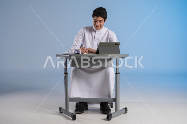 Receiving online lessons via the internet, the concept of back to school, new advanced educational methods, a portrait of a Saudi Arabian Gulf student wearing traditional dress sitting in his seat with gestures of happiness, integrating technology into education in modern schools, blue background