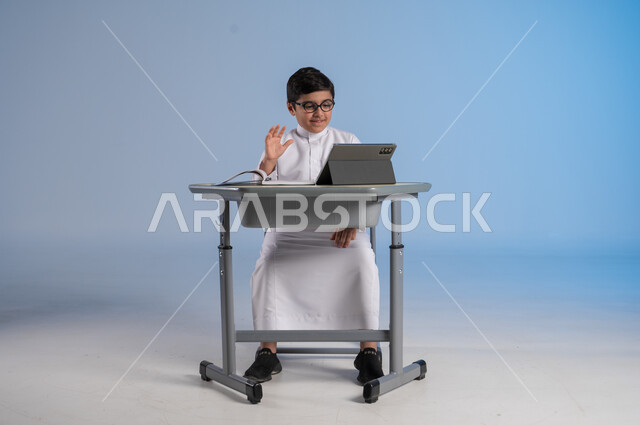 مفهوم العودة إلى المدارس، طرائق تعليمية جديدة متطورة، بورتريه لطالب عربي خليجي سعودي يرتدي الثوب التقليدي يجلس في مقعده بايماءات التركيز والتفاعل، دمج التكنولوجيا بالتعليم في المدارس الحديثة،الاستعداد لتلقي دروس الاون لاين عبر الانترنت، خلفية زرقاء