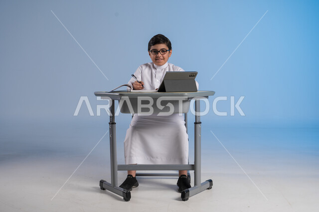 مفهوم العودة إلى المدارس، طرائق تعليمية جديدة متطورة، بورتريه لطالب عربي خليجي سعودي يرتدي الثوب التقليدي يجلس في مقعده بايماءات التركيز والتفاعل، دمج التكنولوجيا بالتعليم في المدارس الحديثة،الاستعداد لتلقي دروس الاون لاين عبر الانترنت، خلفية زرقاء