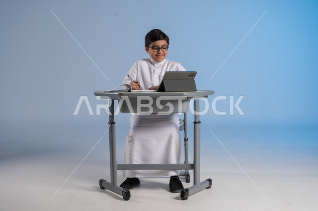 مفهوم العودة إلى المدارس، طرائق تعليمية جديدة متطورة، بورتريه لطالب عربي خليجي سعودي يرتدي الثوب التقليدي يجلس في مقعده بايماءات التركيز والتفاعل، دمج التكنولوجيا بالتعليم في المدارس الحديثة،الاستعداد لتلقي دروس الاون لاين عبر الانترنت، خلفية زرقاء