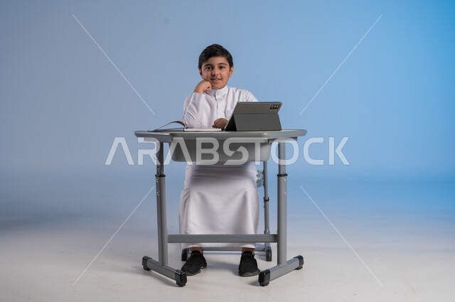 الاستعداد لتلقي دروس الاونلاين عبر جهاز التابلت، مفهوم العودة إلى المدارس، طرائق تعليمية جديدة متطورة، بورتريه لطالب عربي خليجي سعودي يرتدي الثوب التقليدي يجلس في مقعده ينظر الى الكاميرا بتعابير السرور، دمج التكنولوجيا بالتعليم في المدارس الحديثة، خلفية زرقاء