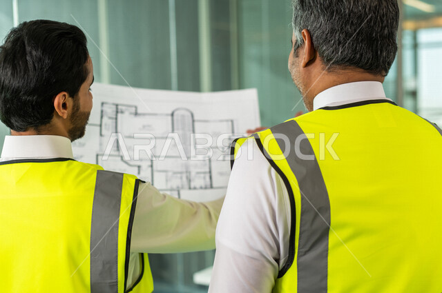 مهندسان معماريان سعوديان خليجيان يحملان مخطط لمشروع البناء للتخطيط وتنفيذ العمل في داخل المكتب في المملكة العربية السعودية, إرتداء سترة الحماية الخاصة بالعمل, تدقيق العمل، الإشراف والنقاش في العمل، مفهوم الهندسة، مهن ووظائف هندسية