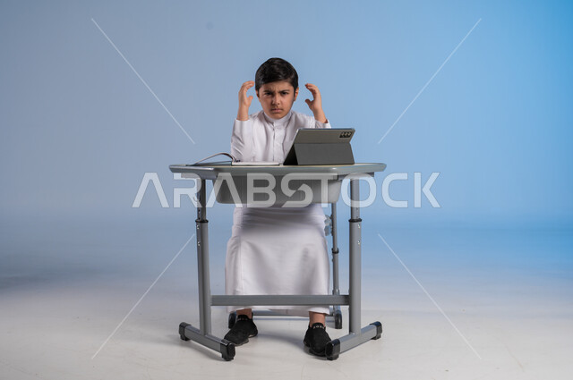 ايماءات اليأس والحزن، مفهوم العودة إلى المدارس، طرائق تعليمية جديدة متطورة، بورتريه لطالب عربي خليجي سعودي يرتدي الثوب التقليدي يجلس في مقعده ينظر الى الكاميرا بايماءات الضجر والتعب من الدراسة، دمج التكنولوجيا بالتعليم في المدارس الحديثة، خلفية زرقاء
