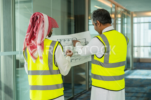 مهندسان معماريان سعوديان خليجيان يحملان مخطط لمشروع البناء للتخطيط وتنفيذ العمل في داخل المكتب في المملكة العربية السعودية, إرتداء سترة الحماية الخاصة بالعمل, تدقيق العمل، الإشراف والنقاش في العمل، مفهوم الهندسة، مهن ووظائف هندسية