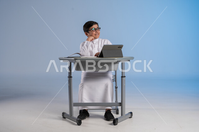 التأمل والتفكير بشيء ما، الاستعداد لتلقي دروس الاونلاين عبر الجهاز اللوحي، طرائق تعليمية جديدة متطورة، بورتريه لطالب عربي خليجي سعودي يرتدي الثوب التقليدي يجلس في مقعده ويحاول النظر لشيء، دمج التكنولوجيا بالتعليم في المدارس الحديثة، خلفية زرقاء