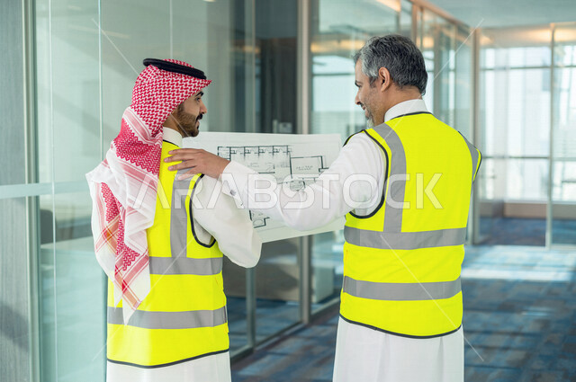 مهندسان معماريان سعوديان خليجيان يحملان مخطط لمشروع البناء للتخطيط وتنفيذ العمل في داخل المكتب في المملكة العربية السعودية, إرتداء سترة الحماية الخاصة بالعمل, تدقيق العمل، الإشراف والنقاش في العمل، مفهوم الهندسة، مهن ووظائف هندسية