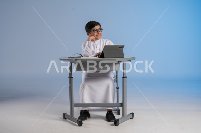 التأمل والتفكير بشيء ما، الاستعداد لتلقي دروس الاونلاين عبر الجهاز اللوحي، طرائق تعليمية جديدة متطورة، بورتريه لطالب عربي خليجي سعودي يرتدي الثوب التقليدي يجلس في مقعده ويحاول النظر لشيء، دمج التكنولوجيا بالتعليم في المدارس الحديثة، خلفية زرقاء