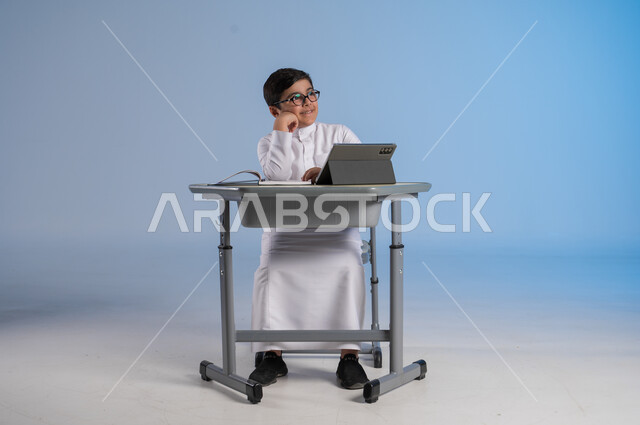 التأمل والتفكير بشيء ما، الاستعداد لتلقي دروس الاونلاين عبر الجهاز اللوحي، طرائق تعليمية جديدة متطورة، بورتريه لطالب عربي خليجي سعودي يرتدي الثوب التقليدي يجلس في مقعده ويحاول النظر لشيء، دمج التكنولوجيا بالتعليم في المدارس الحديثة، خلفية زرقاء