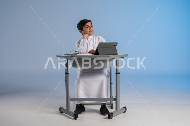 التأمل والتفكير بشيء ما، الاستعداد لتلقي دروس الاونلاين عبر الجهاز اللوحي، طرائق تعليمية جديدة متطورة، بورتريه لطالب عربي خليجي سعودي يرتدي الثوب التقليدي يجلس في مقعده ويحاول النظر لشيء، دمج التكنولوجيا بالتعليم في المدارس الحديثة، خلفية زرقاء