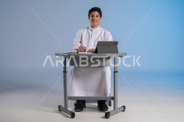 Preparing to receive online lessons via the Internet, new advanced educational methods, portrait of a Saudi Arabian Gulf student wearing traditional dress sitting in his seat while looking at the camera with gestures of pleasure, integrating technology into education in modern schools, blue background
