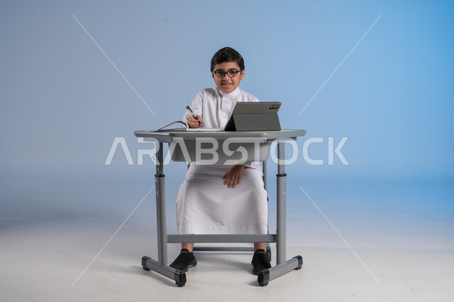 Taking important notes, receiving online lessons via the Internet, new advanced educational methods, portrait of a Saudi Arabian Gulf student wearing traditional dress sitting in his seat with expressions of pleasure in front of him, integrating technology into education in modern schools, blue background