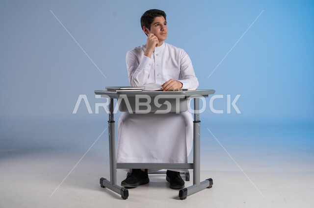 الشغف و السعي نحو النجاح والتفوق, بورتريه لشاب عربي خليجي سعودي يرتدي الثوب التقليدي يجلس في مقعده وينظر إلىشيء ما بايماءات التأمل وشرود الذهن, تحقيق الأحلام والأهداف المستقبلية, خلفية زرقاء
