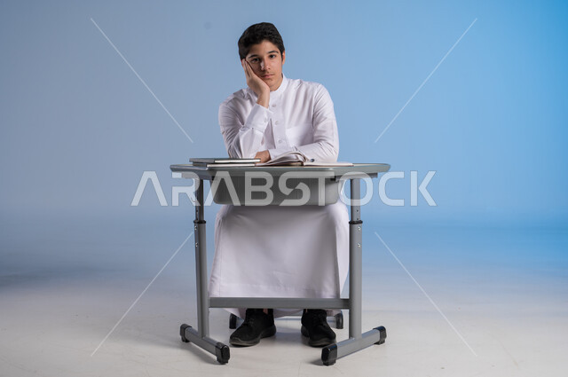 ايماءات الحزن واليأس, بورتريه لطفل عربي خليجي سعودي يرتدي الثوب الأبيض التقليدي يجلس في مقعده وينظر إلى الكاميرا بتعابير البشاشة, تحقيق الأحلام والأهداف المستقبلية, خلفية زرقاء