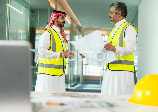 مهندسان معماريان سعوديان خليجيان يحملان مخطط لمشروع البناء للتخطيط وتنفيذ العمل في داخل المكتب في المملكة العربية السعودية, استخدام جهاز اللابتوب، إرتداء سترة الحماية الخاصة بالعمل, تدقيق العمل، الإشراف والنقاش، مهن ووظائف هندسية، فريق عمل خبير