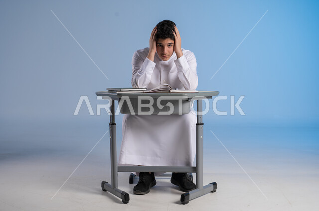 إيماءات العبوس والملل، الشعور والاحساس بالضجر والارهاق من حل الواجبات اليومية، بورتريه شاب عربي خليجي سعودي يرتدي الثوب التقليدي يجلس على مقعده ينظر الى الكاميرا بتعابير التعب، خلفية زرقاء