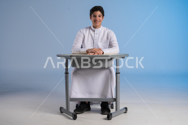 تحقيق الأحلام والأهداف المستقبلية, الشغف و السعي نحو النجاح والتفوق, بورتريه لشاب عربي خليجي سعودي يرتدي الثوب التقليدي يجلس في مقعده وينظر إلى الكاميرا بإيماءات السعادة السرور, خلفية زرقاء