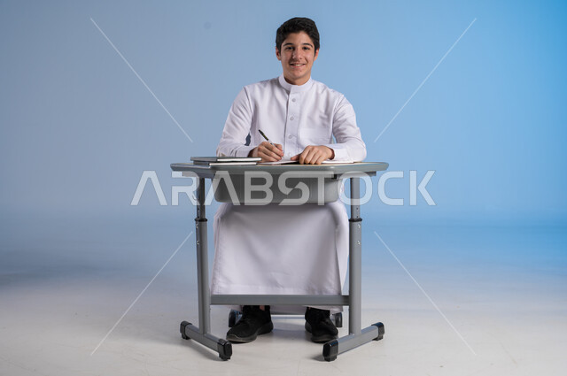 تحقيق الأحلام والأهداف المستقبلية, الشغف و السعي نحو النجاح والتفوق, بورتريه لشاب عربي خليجي سعودي يرتدي الثوب التقليدي يجلس في مقعده وينظر إلى الكاميرا بإيماءات السعادة السرور, خلفية زرقاء