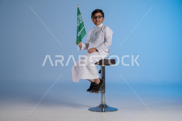 الجلوس على الكرسي والنظر إلى الكاميرا بإيماءات الفرح في يوم العلم  11 مارس، مشاعر الحماس في الاحتفال باليوم الوطني السعودي 23 سبتمبر، بورتريه لولد عربي خليجي سعودي يرتدي الثوب التقليدي يحمل بيده راية المملكة، تقدير وحب الوطن خلفية زرقاء
