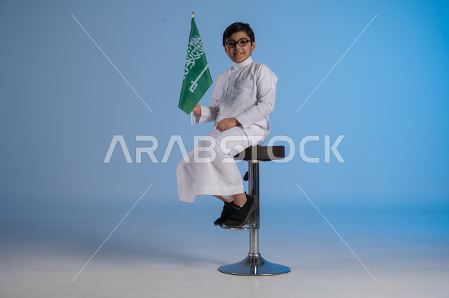 الجلوس على الكرسي والنظر إلى الكاميرا بإيماءات الفرح في يوم العلم  11 مارس، مشاعر الحماس في الاحتفال باليوم الوطني السعودي 23 سبتمبر، بورتريه لولد عربي خليجي سعودي يرتدي الثوب التقليدي يحمل بيده راية المملكة، تقدير وحب الوطن خلفية زرقاء