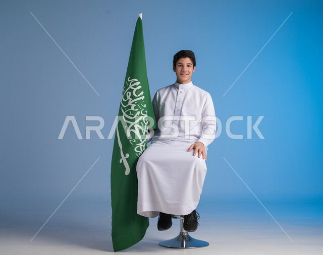 الجلوس على الكرسي والنظر إلى الكاميرا بإيماءات الفرح في يوم العلم  11 مارس، مشاعر الحماس في الاحتفال باليوم الوطني السعودي 23 سبتمبر، بورتريه لشاب عربي خليجي سعودي يرتدي الثوب التقليدي يحمل بيده راية المملكة، خلفية زرقاء