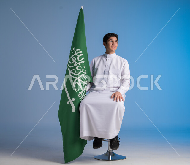 Sitting on chair looking at camera with gestures of joy on Flag Day March 11, feelings of enthusiasm in celebration of Saudi National Day September 23, portrait of a young Saudi Arabian Gulf man wearing traditional dress holding the Kingdom's flag in his hand, blue background