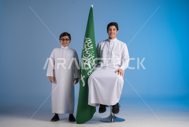 تقدير وحب الوطن، النظر إلى الكاميرا بإيماءات الفرح في يوم العلم  11 مارس، مشاعر الحماس في الاحتفال باليوم الوطني السعودي 23 سبتمبر، بورتريه لأخين عربيين خليجيين سعوديين يرتديان الثوب التقليدي يقفان بجانب راية المملكة، خلفية زرقاء