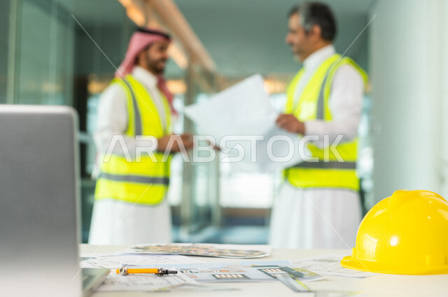 مهندسان معماريان سعوديان خليجيان يحملان مخطط لمشروع البناء للتخطيط وتنفيذ العمل في داخل المكتب في المملكة العربية السعودية, استخدام جهاز اللابتوب، إرتداء سترة الحماية الخاصة بالعمل, تدقيق العمل، الإشراف والنقاش، مهن ووظائف هندسية، فريق عمل خبير
