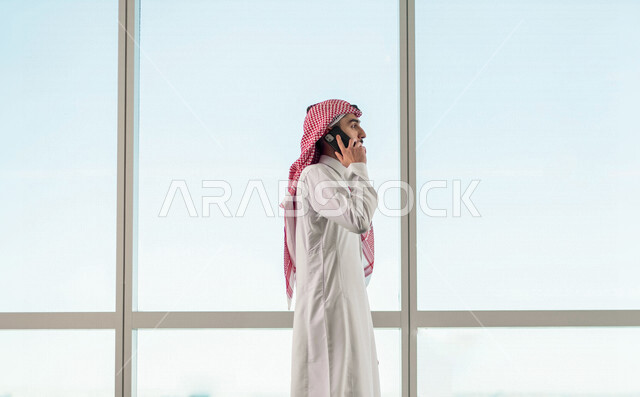 A Saudi Arabian Gulf man in the waiting room, making some calls through a mobile phone, preparing to travel on a business trip, waiting hall at the airport, tourism and travel