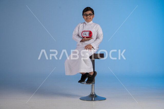 التنبيهات والتذكيرات الثابته، أهمية الدقة والالتزام في المواعيد، بورتريه لفتى عربي خليجي سعودي  يرتدي الثوب التقليدي ينظر الى الكاميرا يحمل بيده ساعة رقمية, إنجاز المهامات في الوقت، خلفية زرقاء