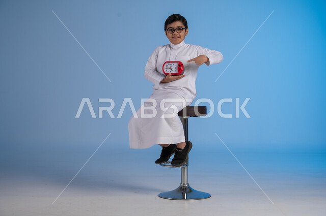 Fixed alerts and reminders, the importance of accuracy and commitment to appointments, portrait of a Saudi Arabian Gulf boy wearing traditional dress looking at the camera holding a digital watch, completing tasks on time, blue background