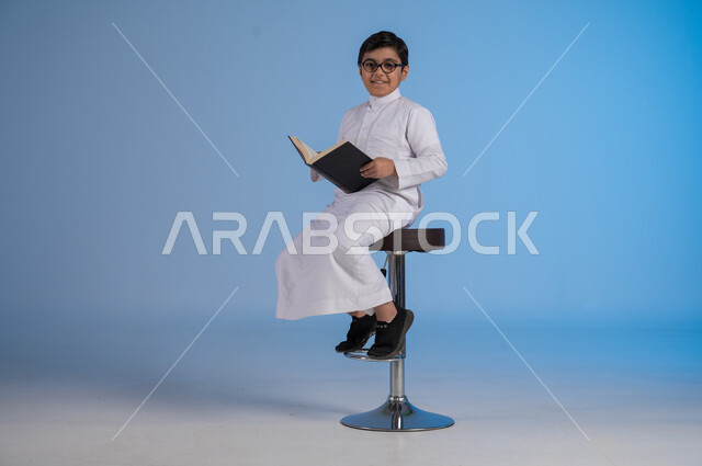 متابعة الدروس والواجبات، تطوير المناهج التعليمية في السعودية, بورتريه لولد عربي سعودي خليجي يرتدي الثوب التقليدي والنظارات الطبية يجلس على كرسي يحمل الكتاب المدرسي, بداية العام الدراسي الجديد, إيماءات الاجتهاد وتحضير الدروس, خلفية زرقاء