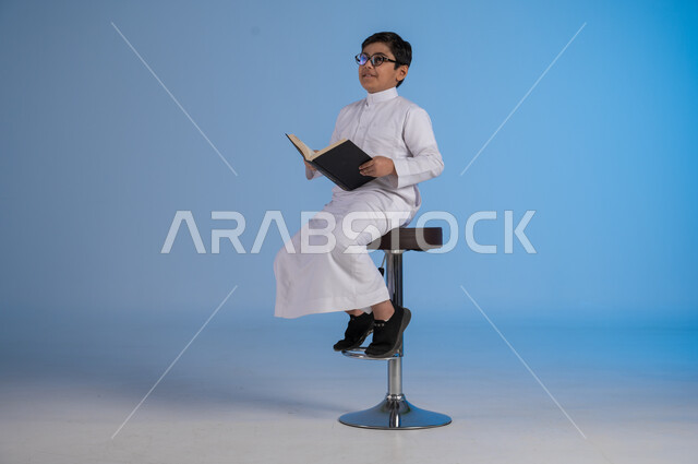 Follow up lessons and assignments, developing educational curricula in Saudi Arabia, portrait of a Saudi Arabian Gulf boy wearing traditional dress and glasses sitting on a chair holding a school book, beginning of the new school year, gestures of diligence and preparing lessons, blue background