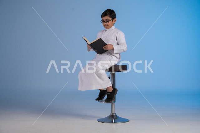 متابعة الدروس والواجبات، تطوير المناهج التعليمية في السعودية, بورتريه لولد عربي سعودي خليجي يرتدي الثوب التقليدي والنظارات الطبية يجلس على كرسي يحمل الكتاب المدرسي, بداية العام الدراسي الجديد, إيماءات الاجتهاد وتحضير الدروس, خلفية زرقاء