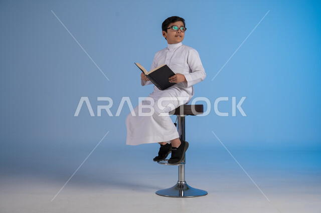 Follow up lessons and assignments, developing educational curricula in Saudi Arabia, portrait of a Saudi Arabian Gulf boy wearing traditional dress and glasses sitting on a chair holding a school book, beginning of the new school year, gestures of diligence and preparing lessons, blue background
