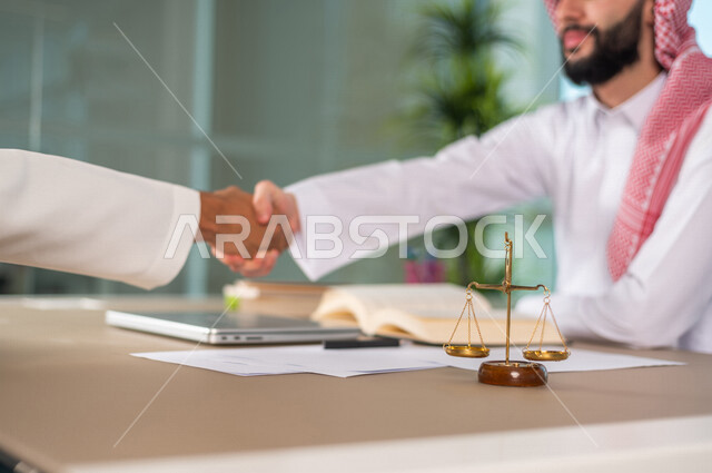 رجل عربي خليجي سعودي يعمل في مجال القضاء، امامه ميزان العدل، استلام قضية جديدة، يدرس اوراق القضية، يصافح احد موكليه