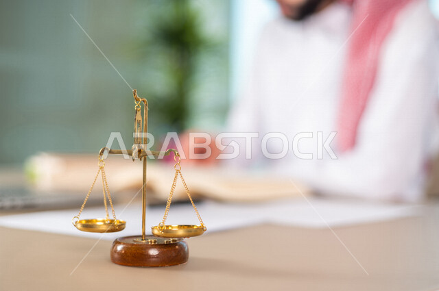 A blurry picture of a Saudi Arabian Gulf man working in the field of justice, wearing traditional Saudi dress, in front of him is the scale of justice, studying case papers, using a computer to complete tasks