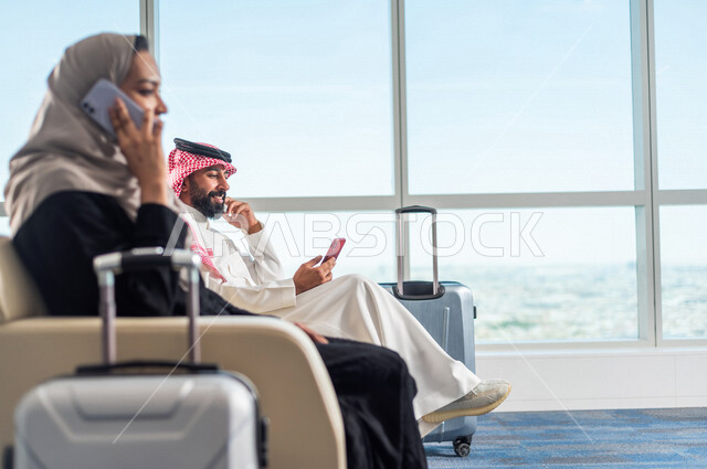 رجل عربي خليجي سعودي يتصفح الانترنت من خلال الهاتف المحمول وبجانبه مراة خليجية تجري الاتصالات، صالة الانتظار في المطار، سياحة وسفر، قضاء أوقات ممتعة